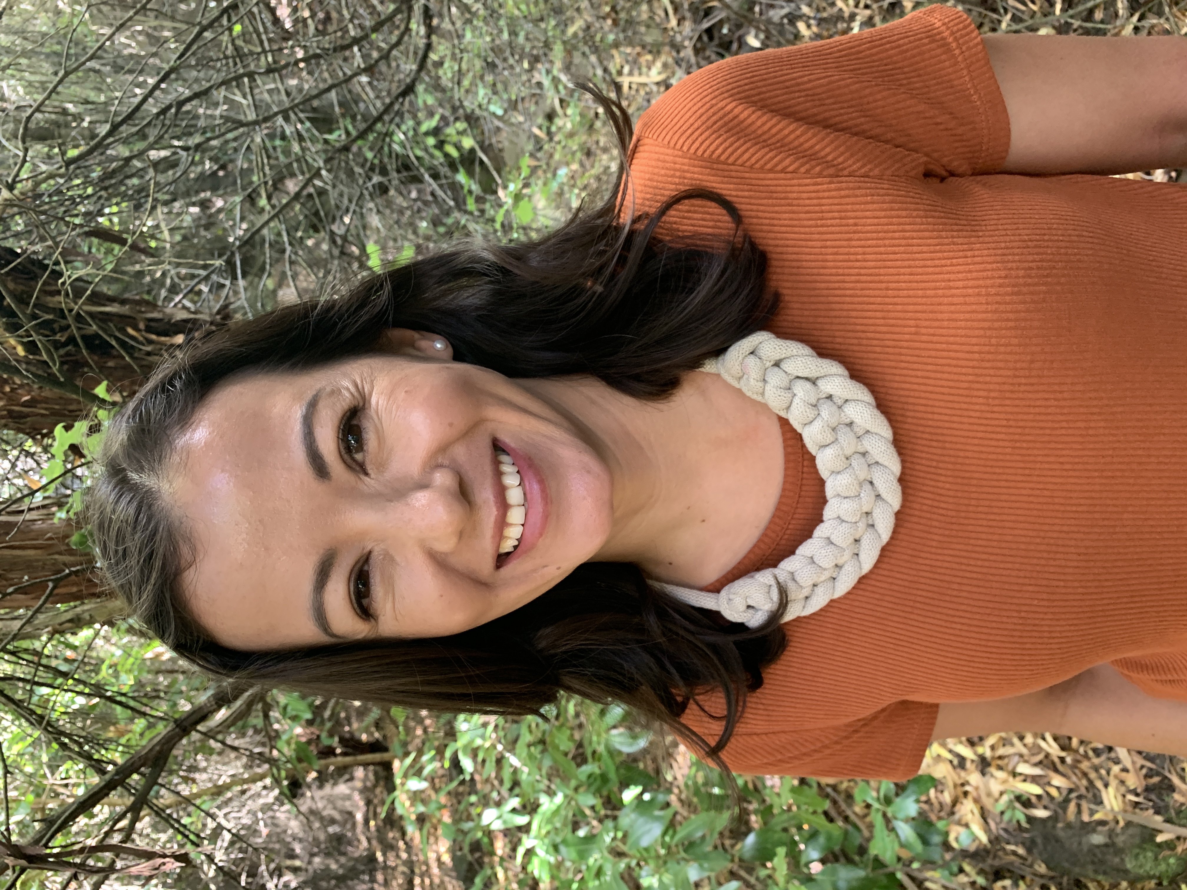 A photo of me in the bush, from above the waist, smilling at the camera,  wearing an  orange top and woven necklace.